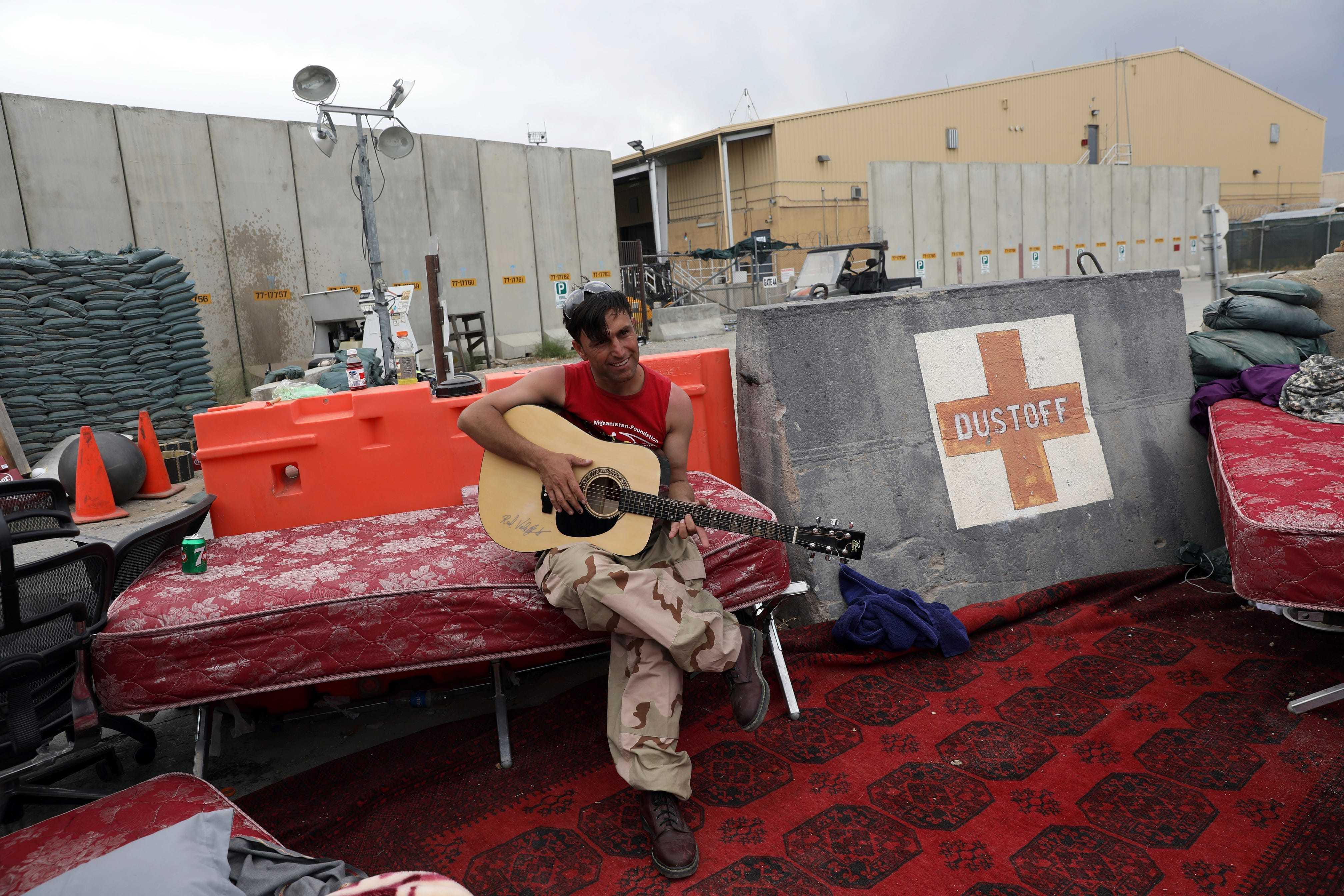 An Afghan soldier sitting on a red mattress plays a guitar.