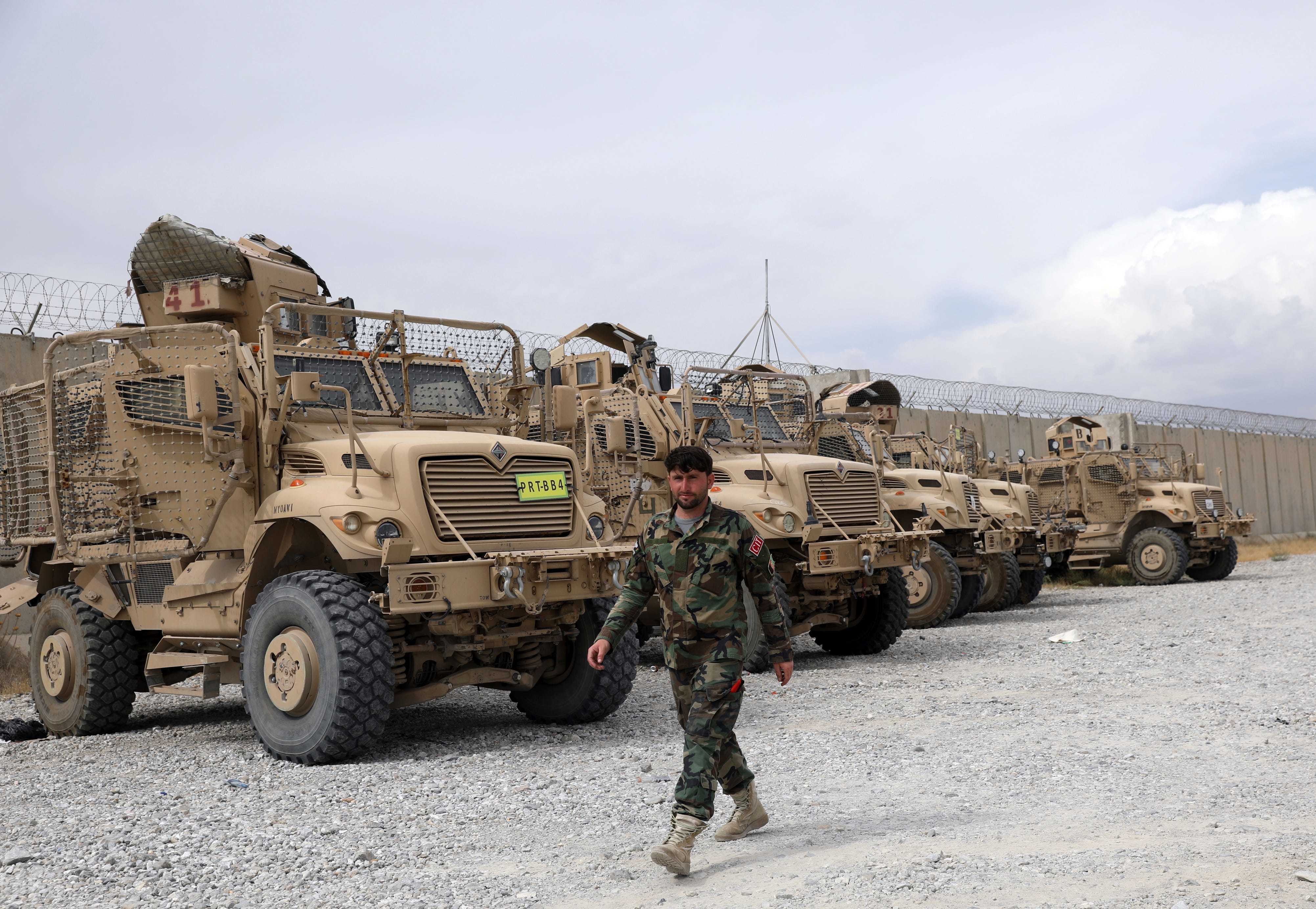 An Afghan soldier walks past several large beige army vehicles.