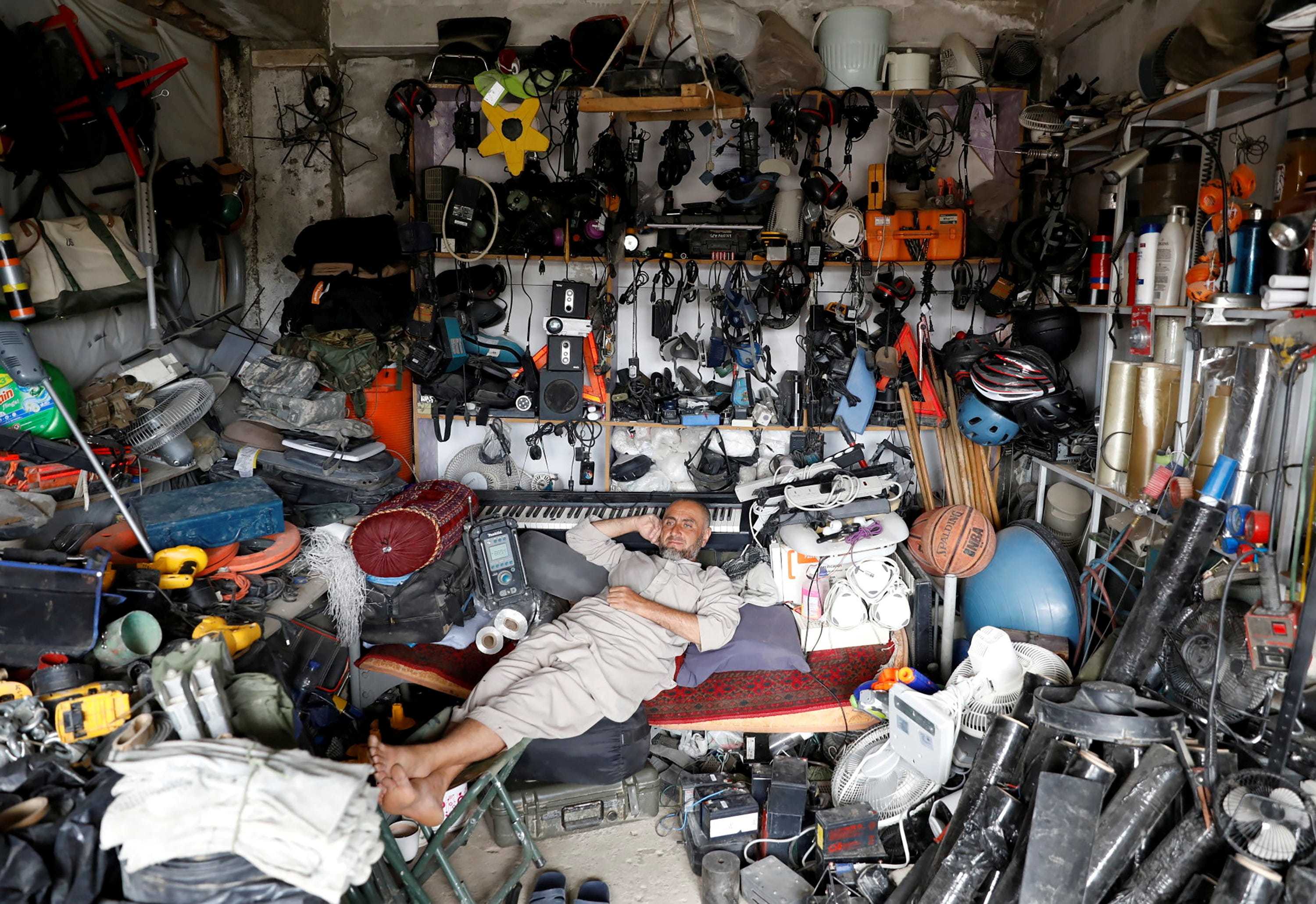 An Afghan man rests on a couch in his shop surrounded by items on the floor and the walls.