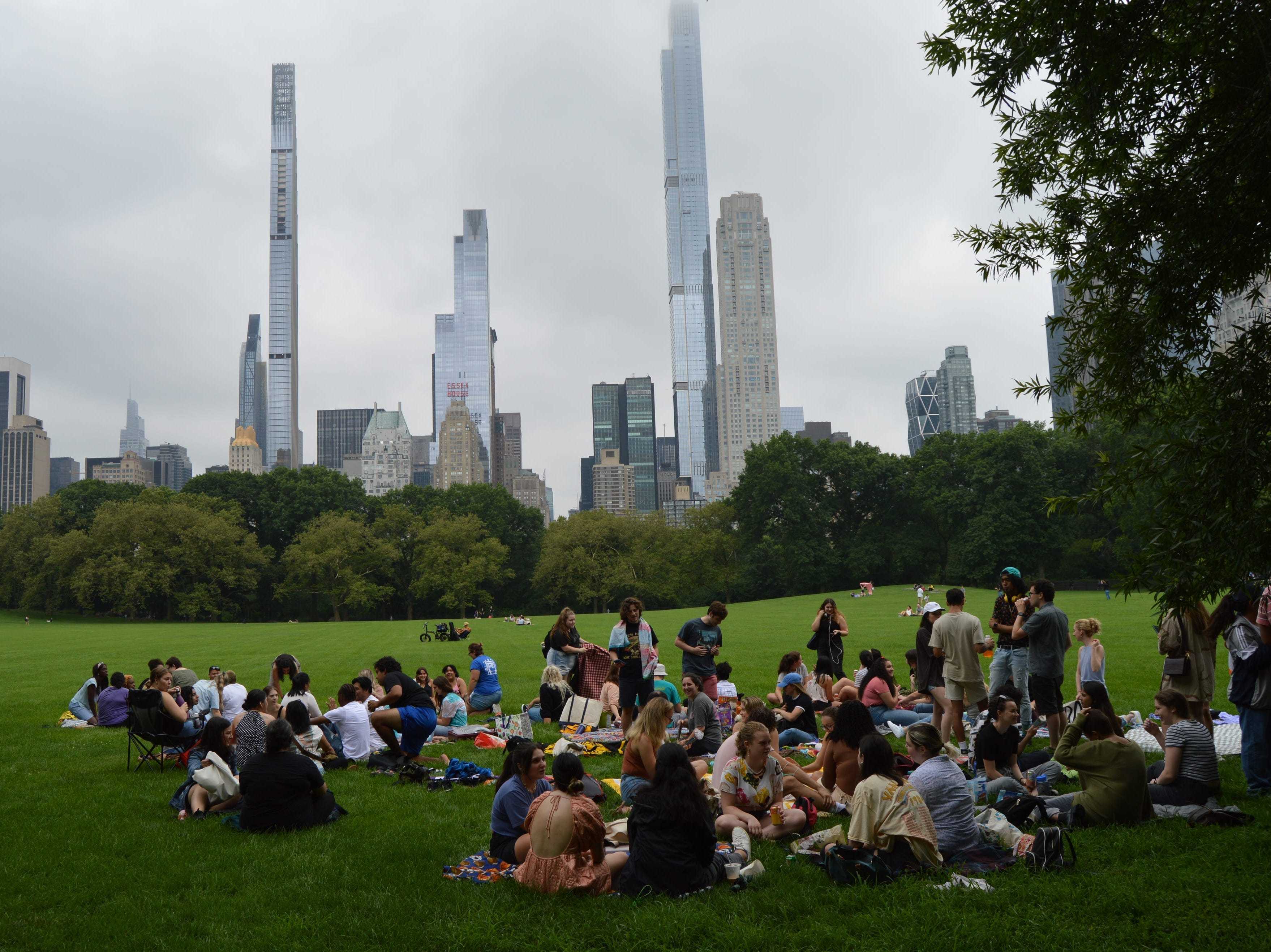 Dozens of people sit on the grass chatting in Central Park, with New York City skyscrapers behind them.