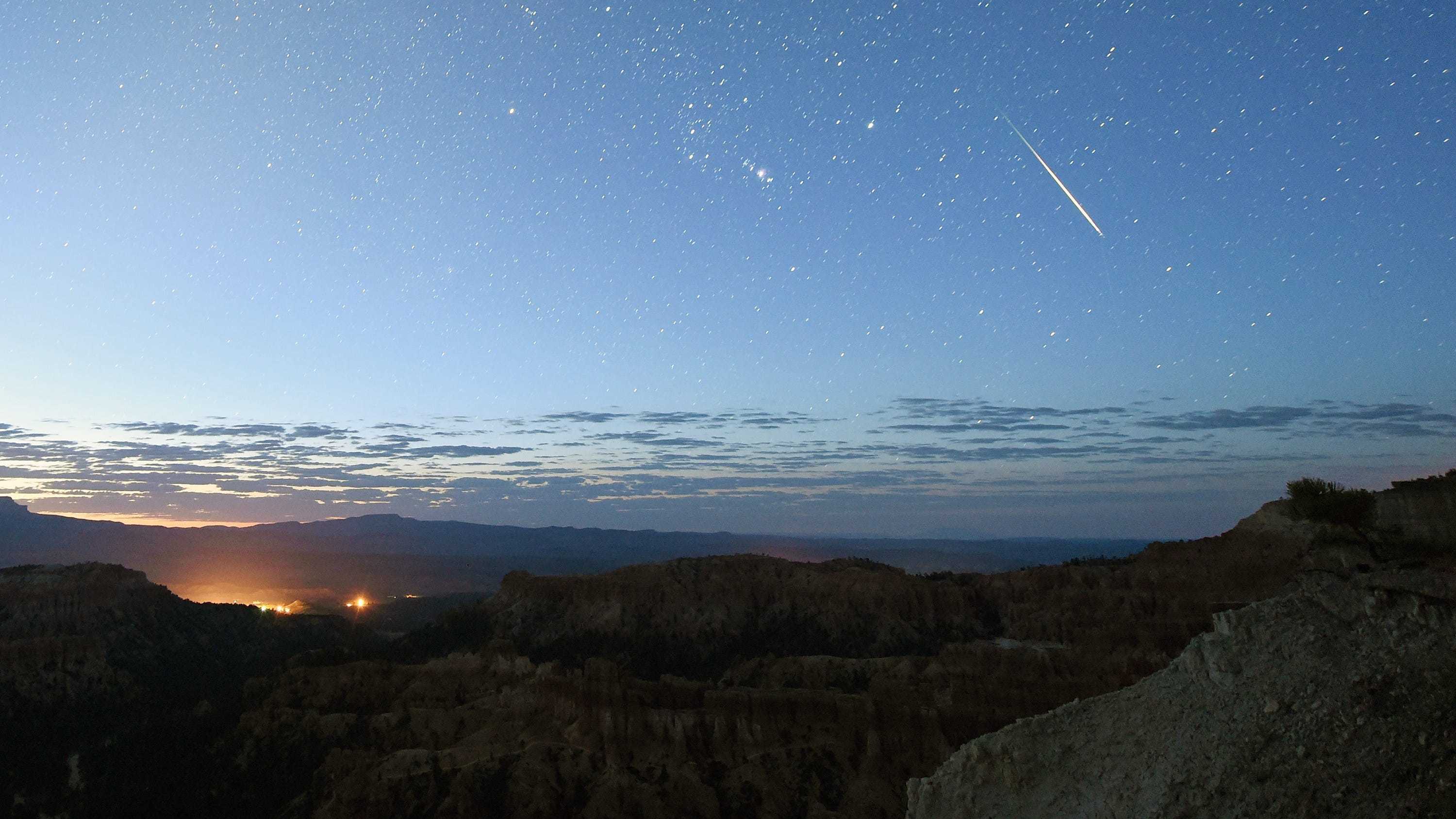 A meteor streaking across a small section of blue sky with stars and the tip of the sun in the lower-left corner visible.