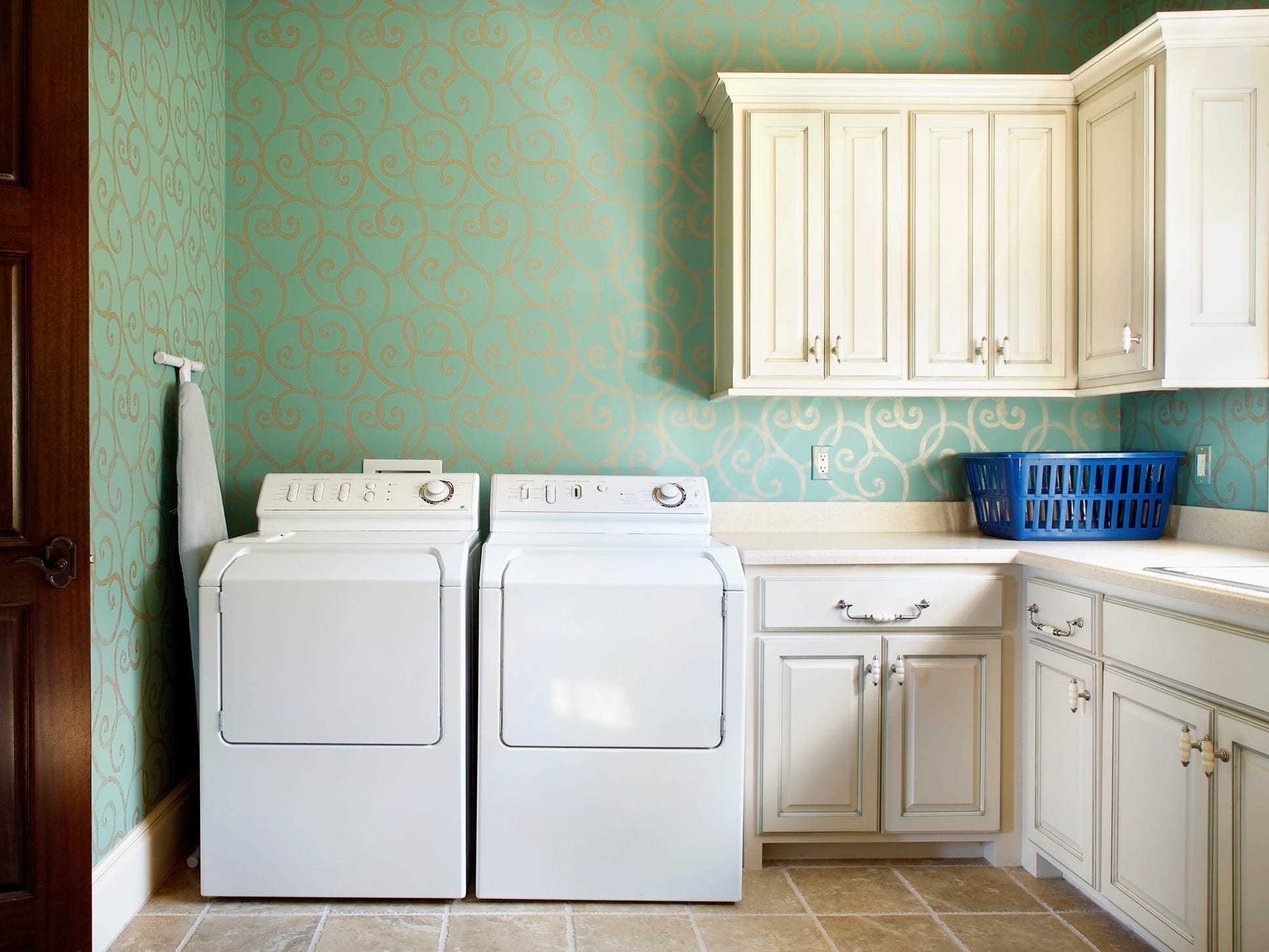 Wide shot of a laundry room with green and gold wallpaper and white cabinets.