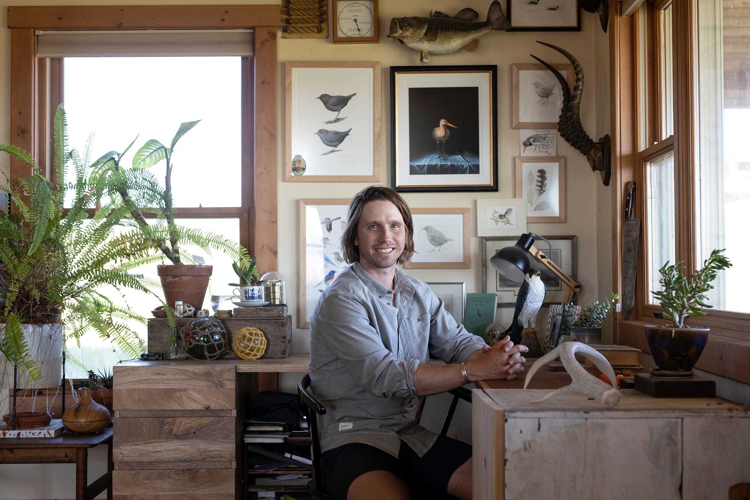 Ecologist Charles Post wears a gray button-up shirt while sitting in an office decorated with plants and pictures of birds.