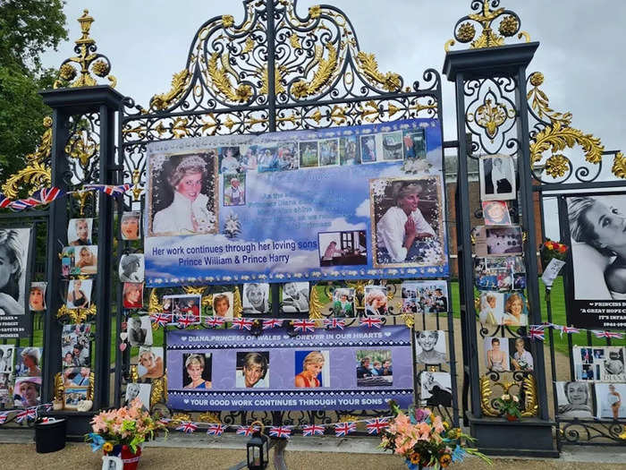 The palace gates are lined with banners, photos of the royal, Union Jack flags, and flowers.