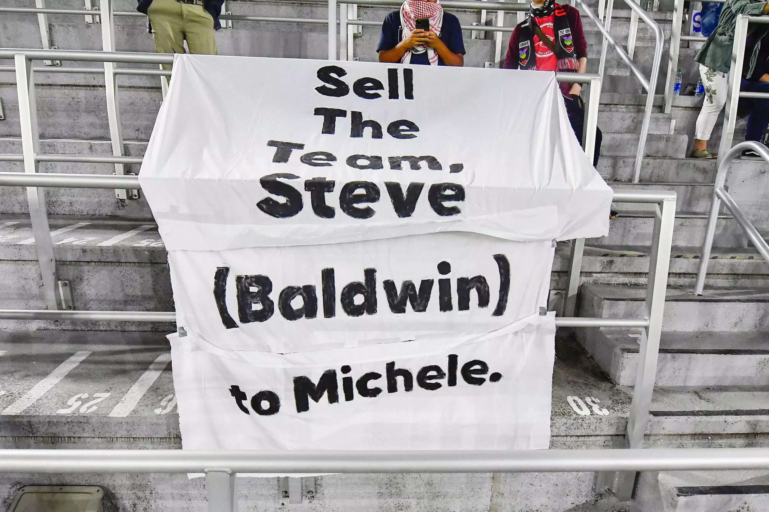 Fans hold signs lobbying Washington Spirit owner Steve Baldwin to sell his stake in the NWSL franchise.