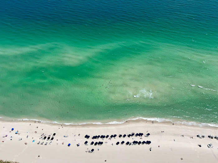 And a white sand beach speckled with umbrellas and sunbathers, resembling a Gray Malin photo.