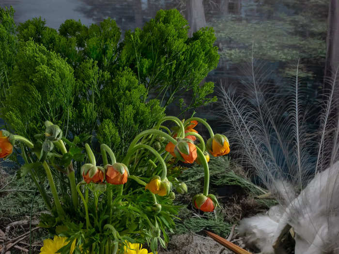 The background has birds on a forest landscape. The foreground has a white cloth with a vase full of wilted flowers and greens.