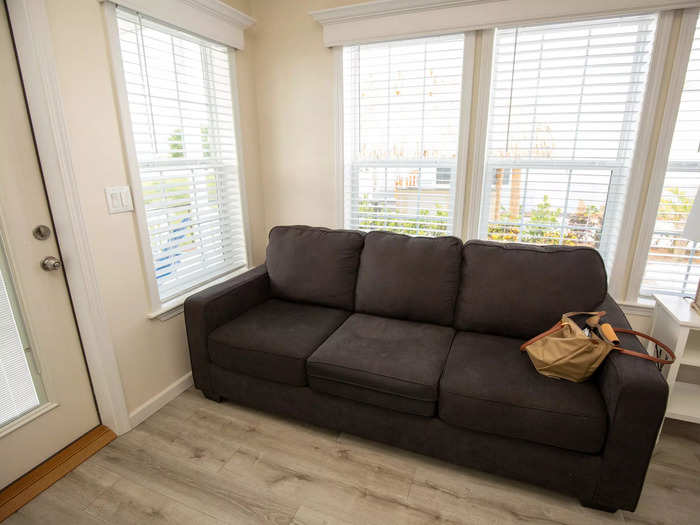 The living room has large windows, a wall-mounted television, a sleeper couch, and a narrow table with several seats.