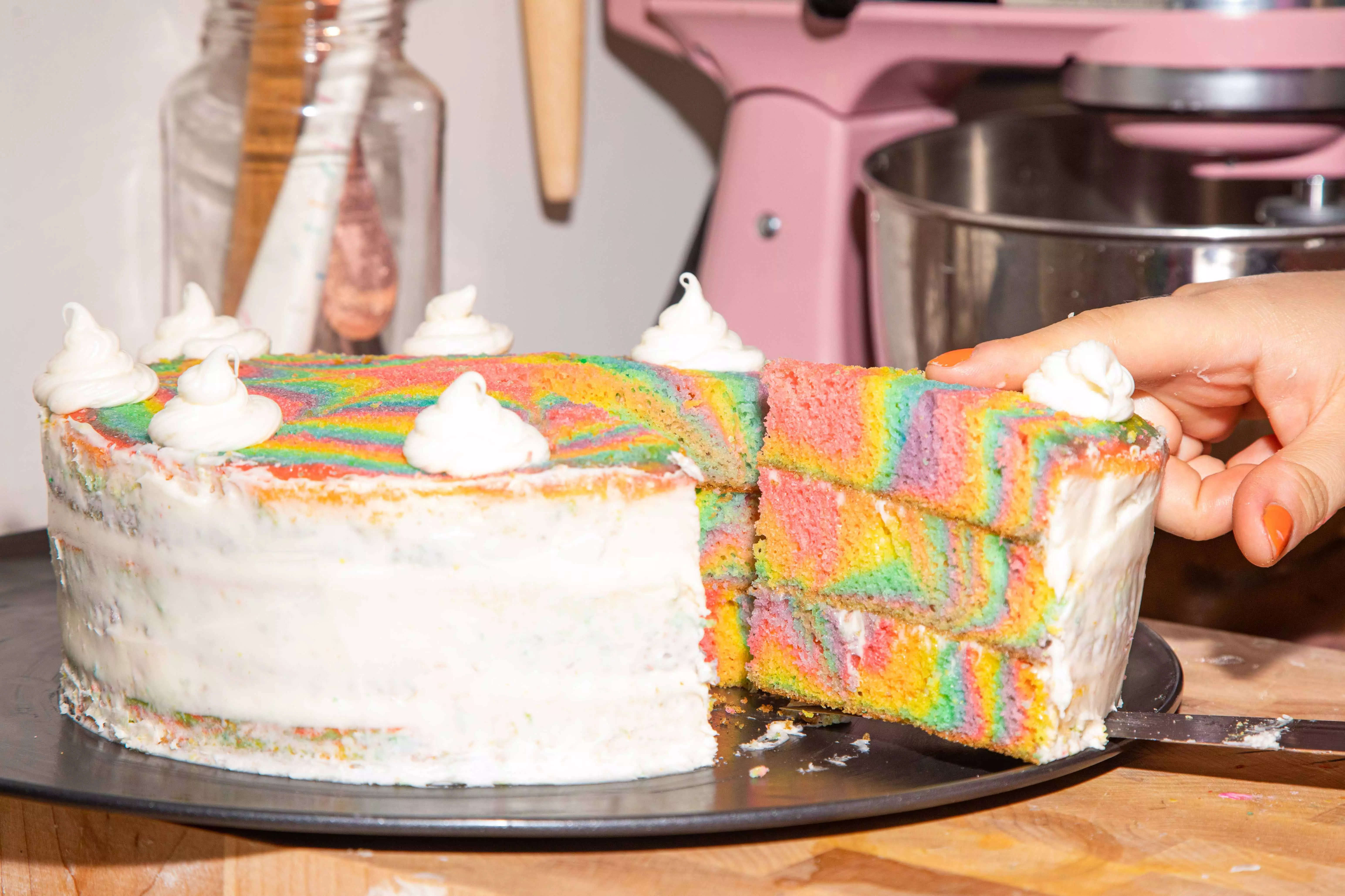 A slice of tie-dye patterned rainbow cake being cut out of a whole cake
