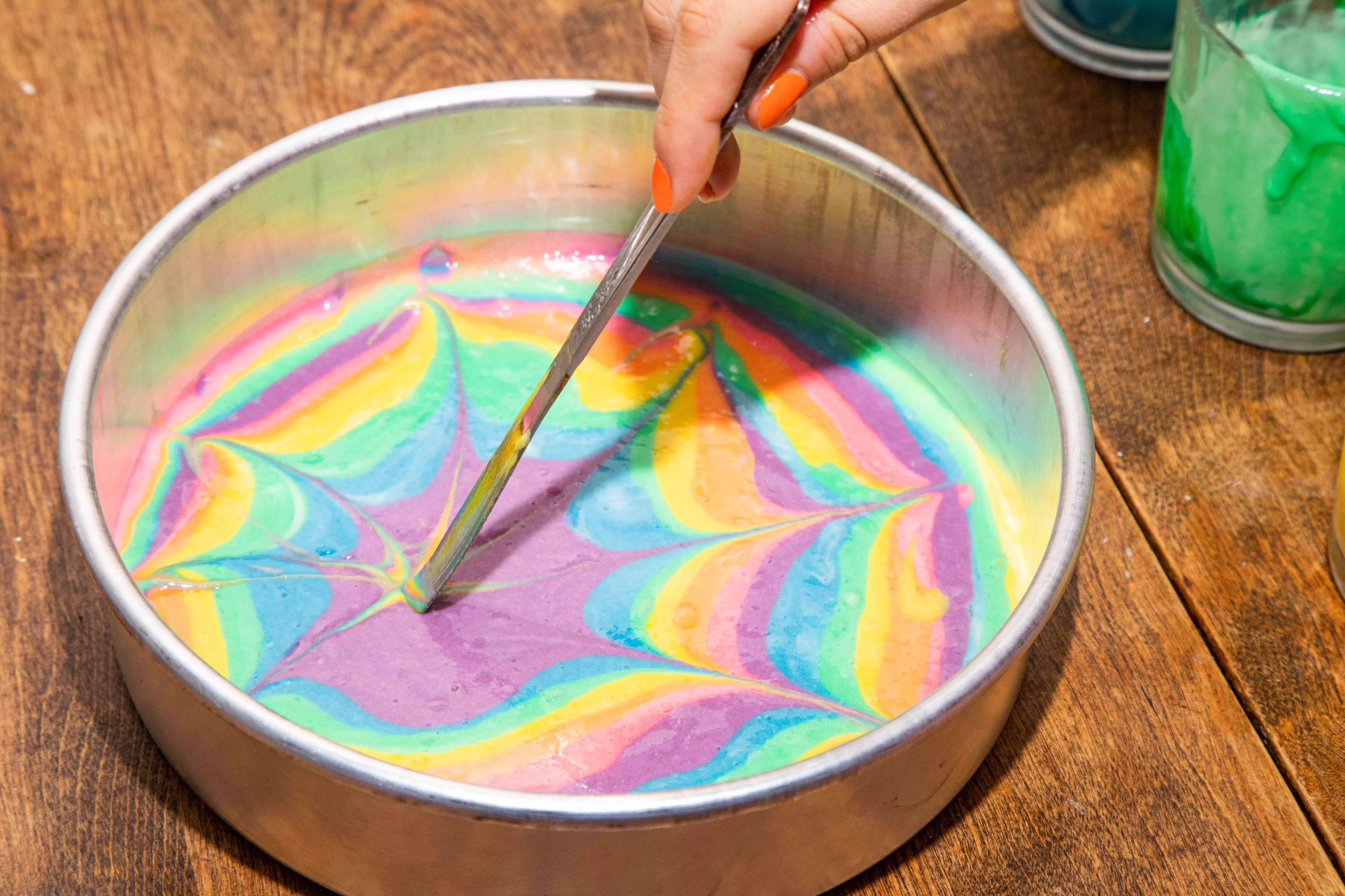 A hand holding a butter knife, using it to swirl tie-dye cake batter in a cake pan