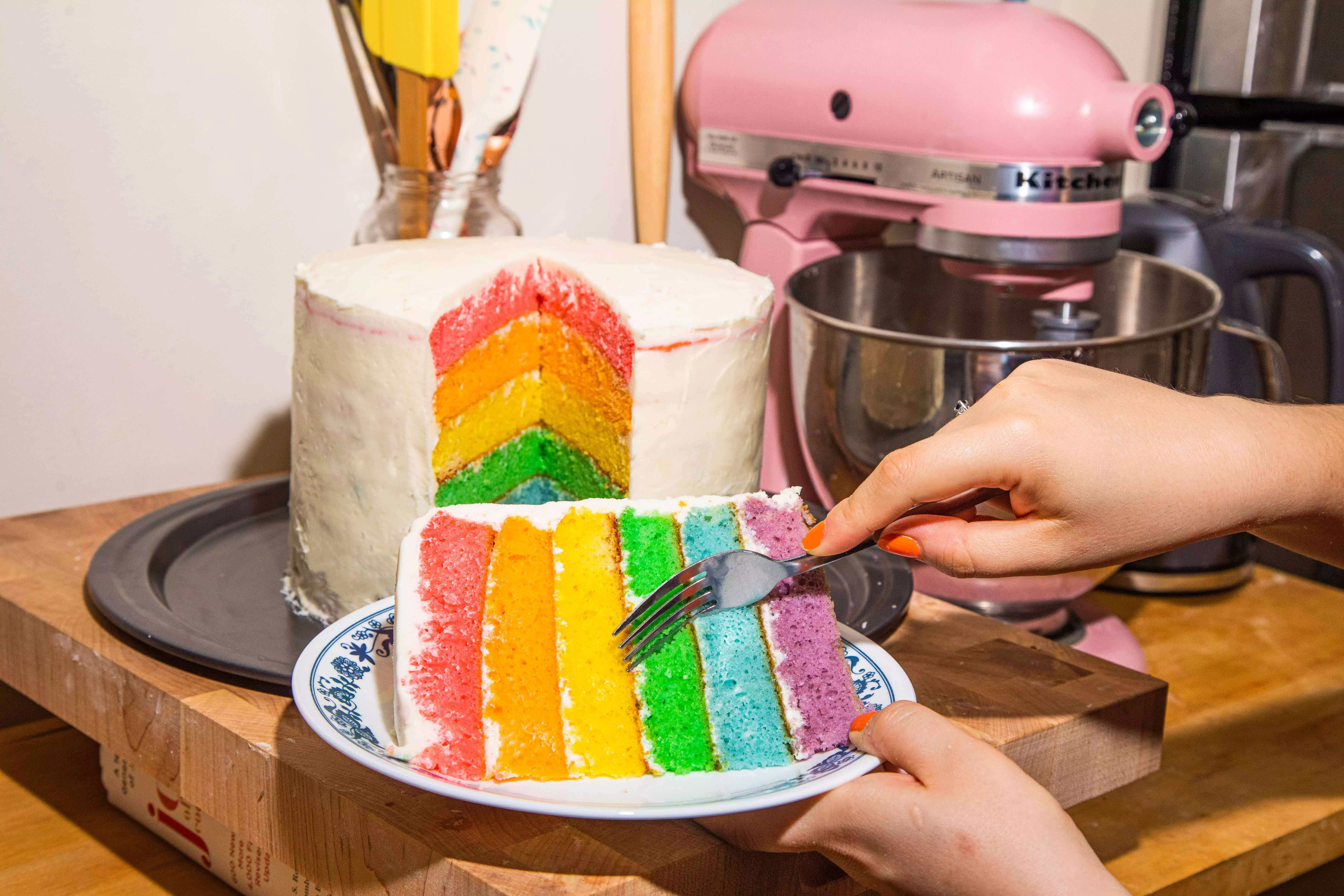 A hand holding a fork cutting into a slice of a layered rainbow cake, with the whole cake in the background