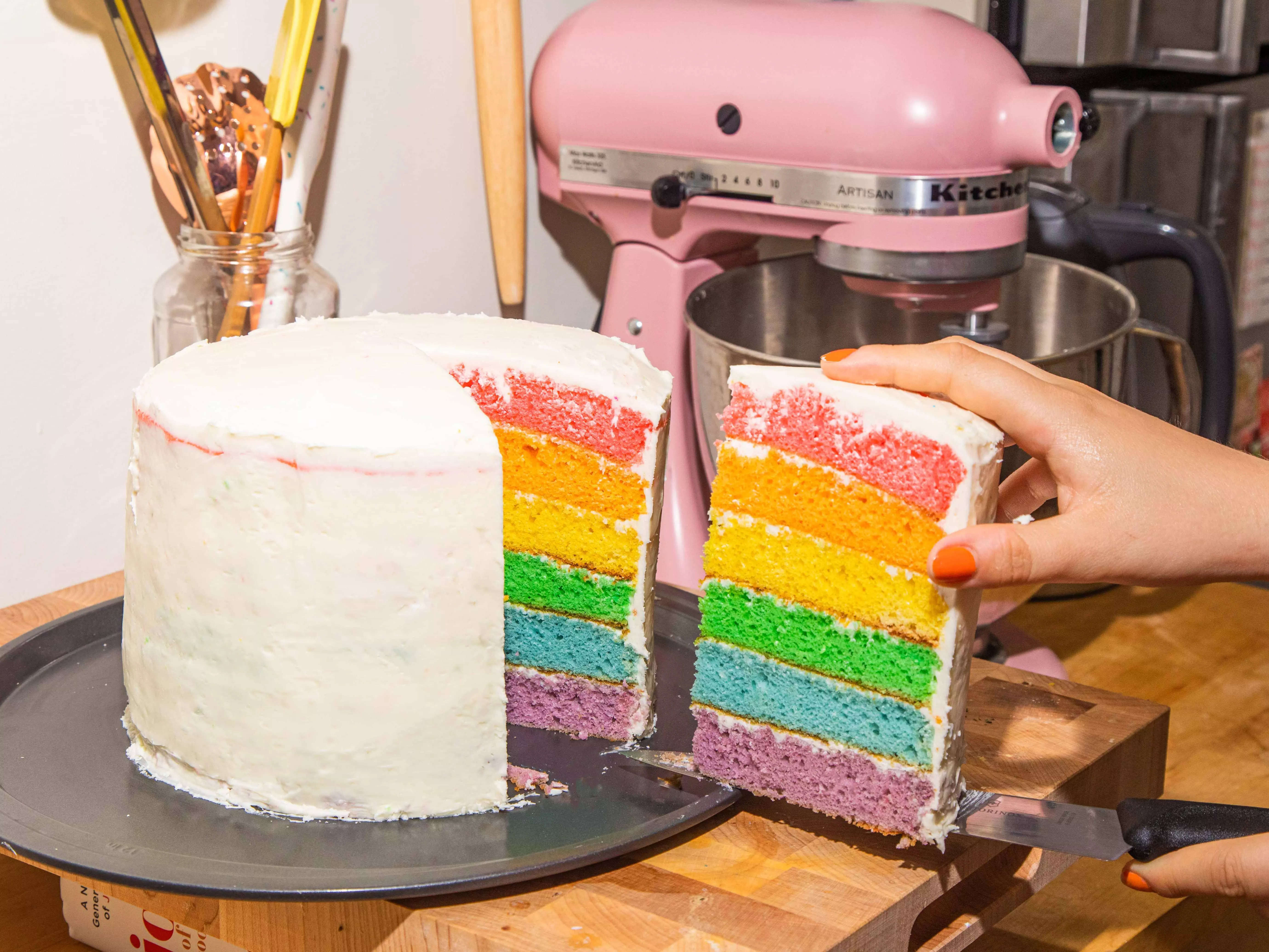 A hand holding a slice of a six layer rainbow cake with the full cake behind it