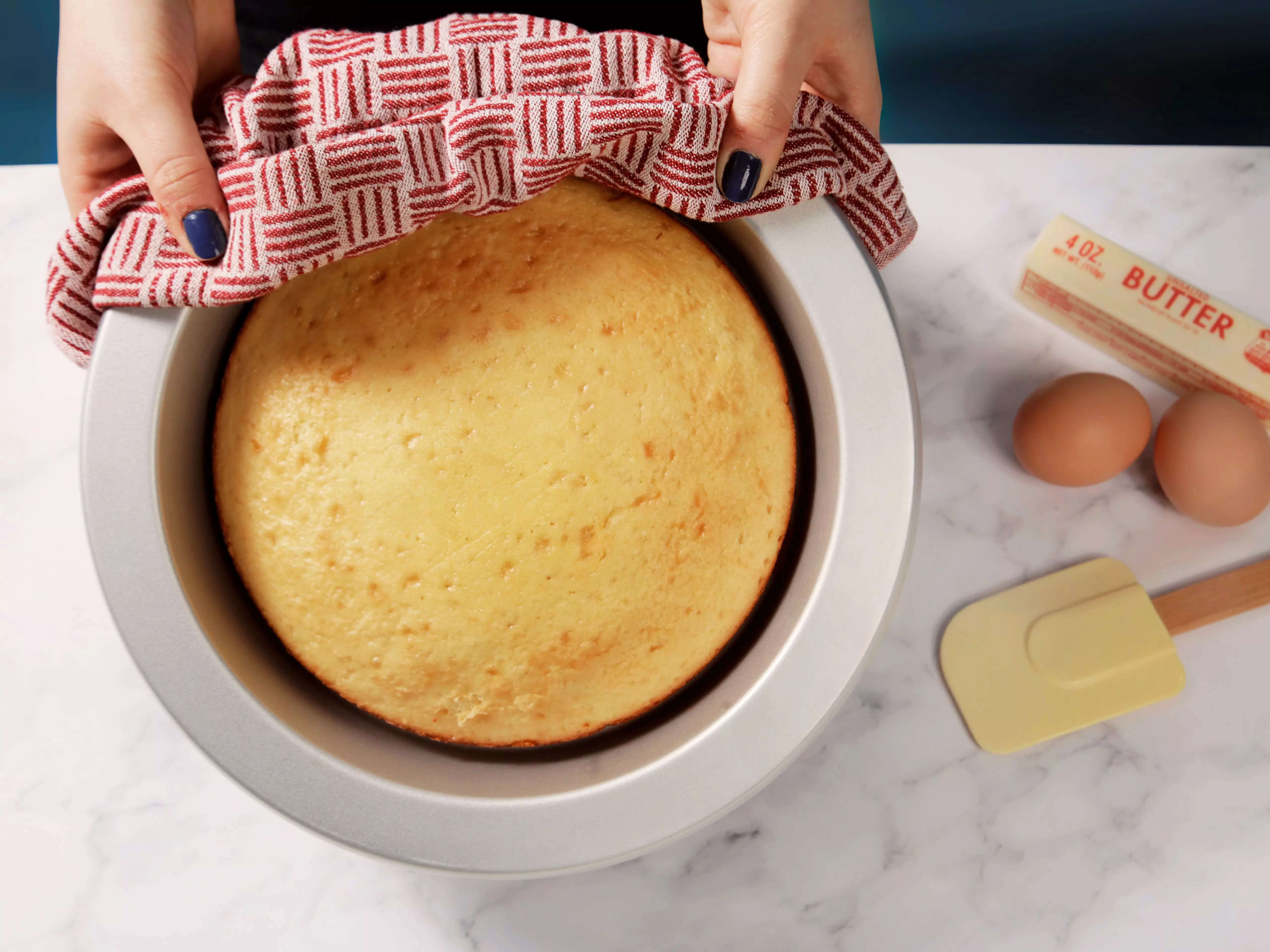 Hands holding a KitchenAid cake pan with a baked cake inside next to a yellow spatula, two eggs, and a stick of butter - best cake pans in 2022