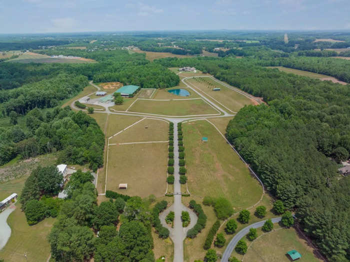 In this aerial view, the main house is in the back-right, the covered horse arena is off to the left, and the guest house is in the lower-left area partially hidden by trees.