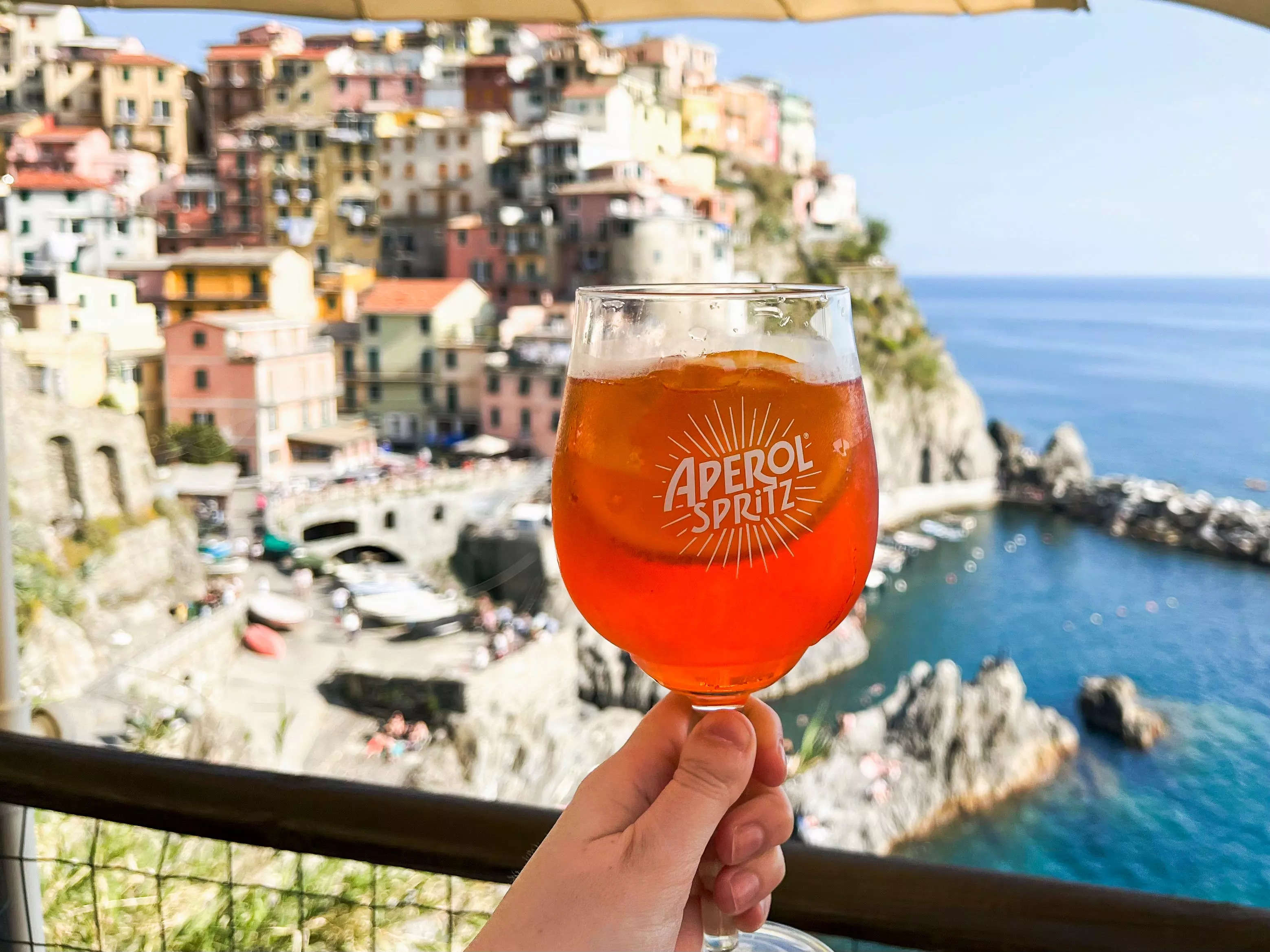 The author holds an Aperol Spritz in front of a colorful Italian town by the water.