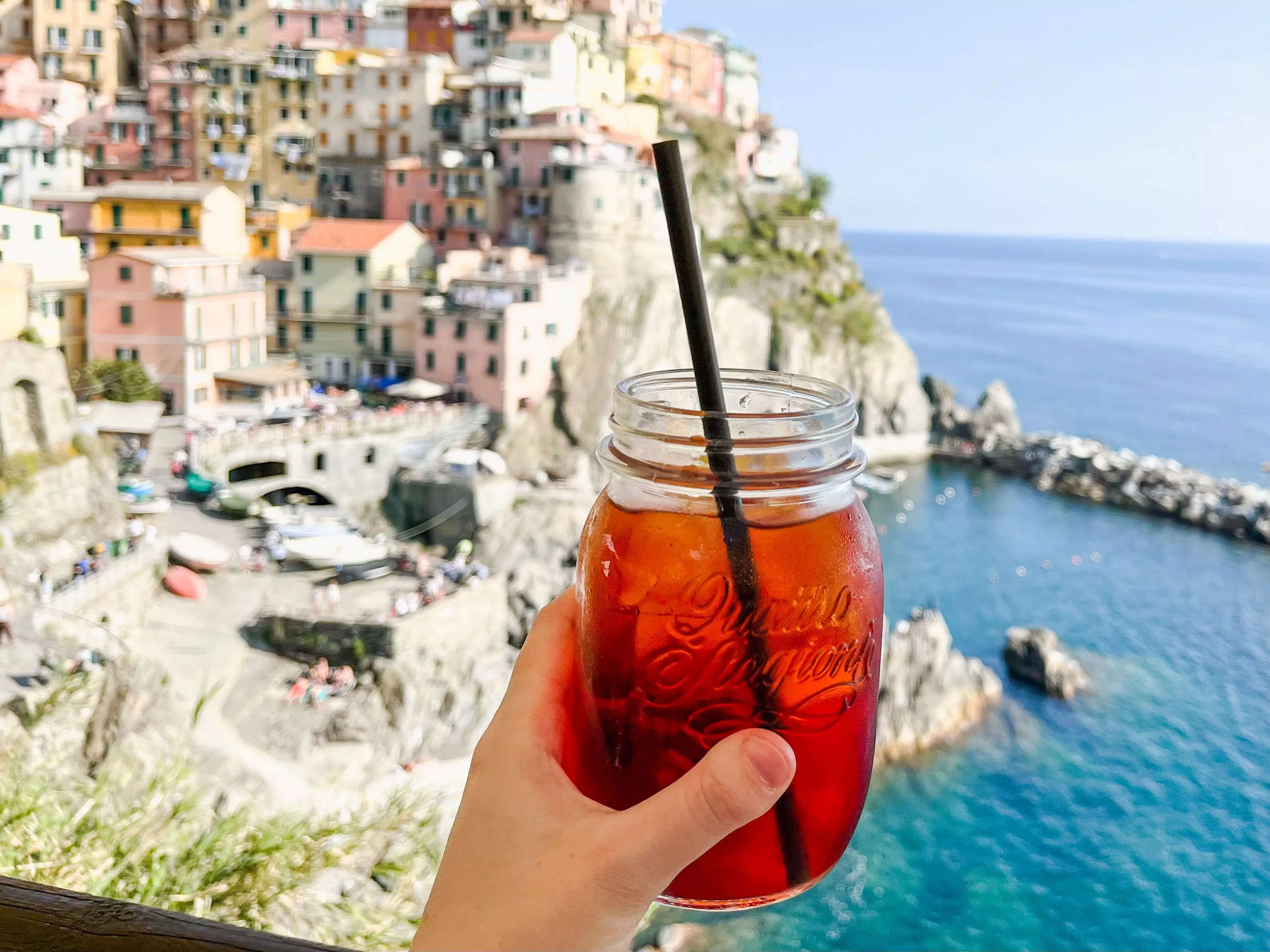 The author holds an Americano cocktail in front of a colorful Italian town by the water.