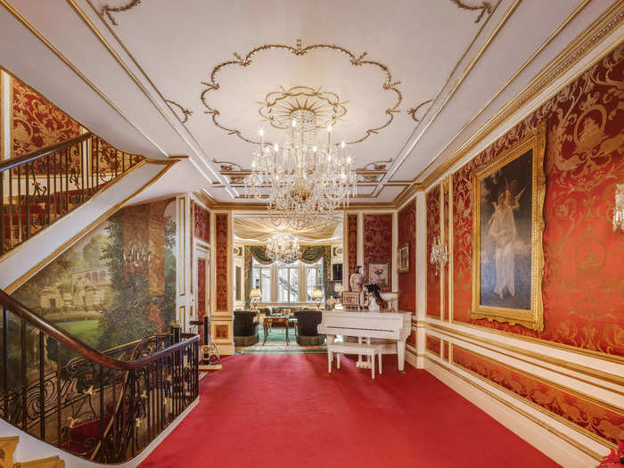 One of the hallways of the home includes a crystal chandelier and a white grand piano.