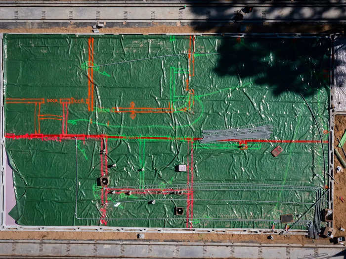 Next, a floor plan is painted on top. The red lines in this picture are where the walls will be built. The tube-like structures protruding out of the ground mark where plumbing and electrical hookups have been placed within the floor plan.