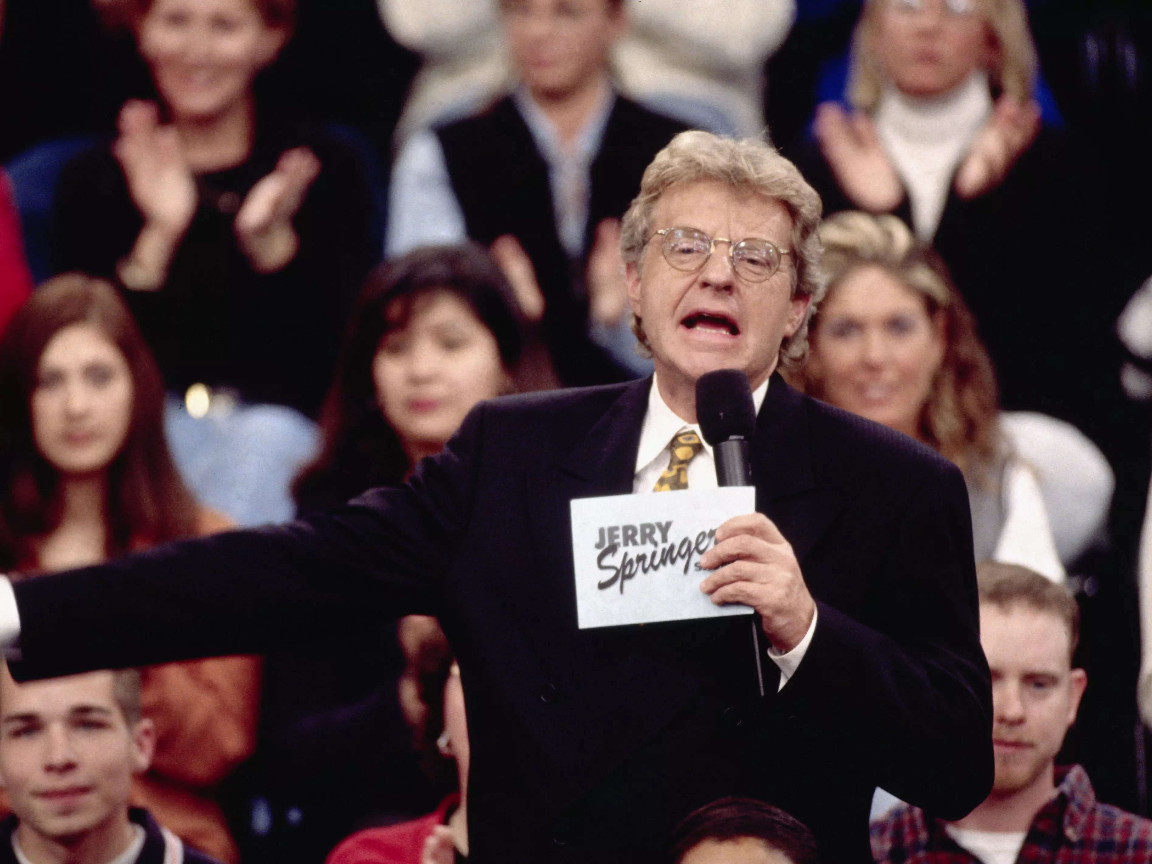 Jerry Springer speaking into a microphone and gesturing to the audience behind him on the set of "The Jerry Springer Show."
