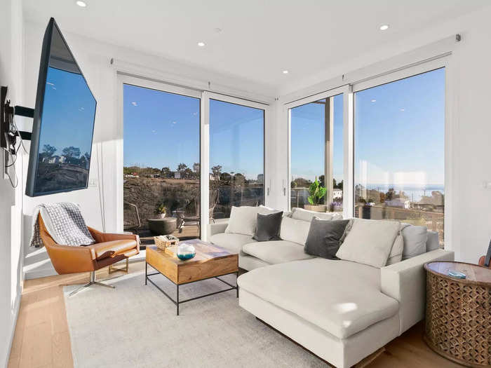 The great room has a living area, a dining area, and a kitchen, with massive windows that frame cliff and ocean views and let in California sunshine.